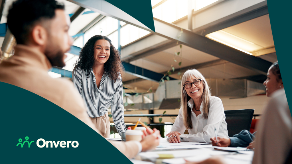 A diverse group of people in a large office environment are around a table, engaged in lively conversation and laughter.